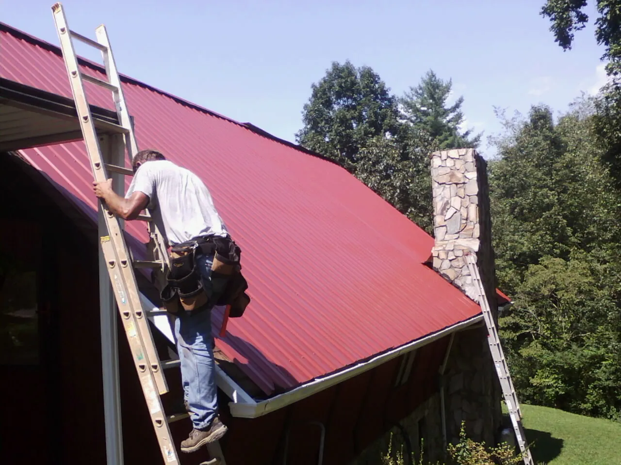 Roofer on a ladder beside a red metal roof and stone chimney — Soffit & Fascia Repair by Roofing Brothers in Lake Charles